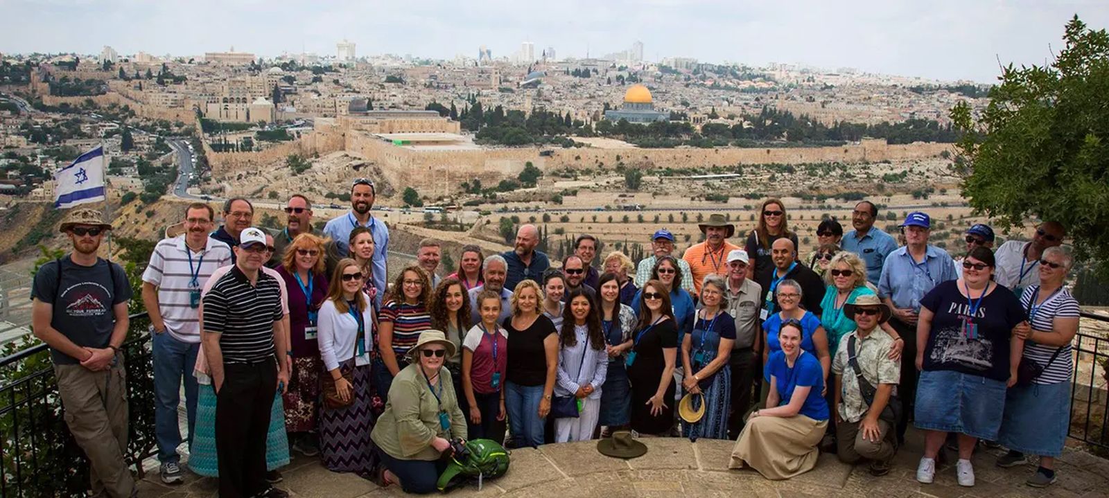 A large Christian group tour gathered at the Mount of Olives overlook, with Jerusalem's Old City, the Dome of the Rock and an Israeli flag behind them